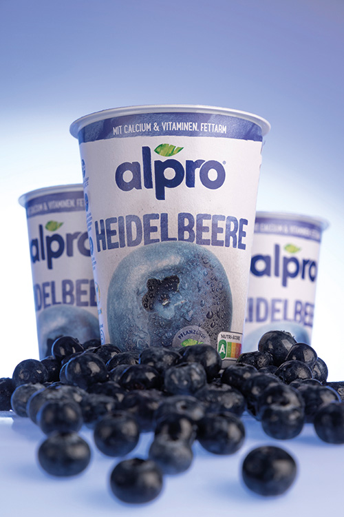 Ein Produktfoto von Alpro Heidelbeer-Joghurt. Drei Stück stehen im Zentrum des Bildes, der vordere thront auf Blaubeeren.